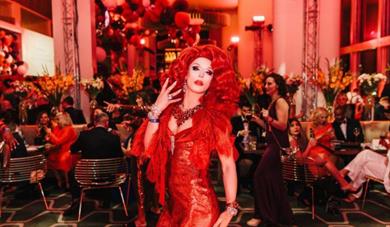 a drag queen in a red dress stood in a function room