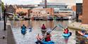 A group of people kayaking on the canal wearing brightly coloured buoyancy aids. They have their backs to the camera and are paddling into Gas Street