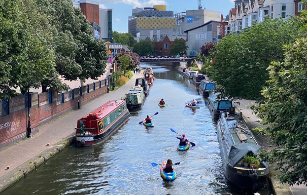 People paddle away from the camera along the canal. Brightly coloured canal boats are moored at the edges and Birmingham Library can be seen in the di