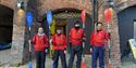 A group of four people pose with their kayak paddles in front of the Roundhouse. They are wearing bright red buoyancy aids and waterproof trousers and