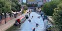 People paddle away from the camera along the canal. Brightly coloured canal boats are moored at the edges and Birmingham Library can be seen in the di