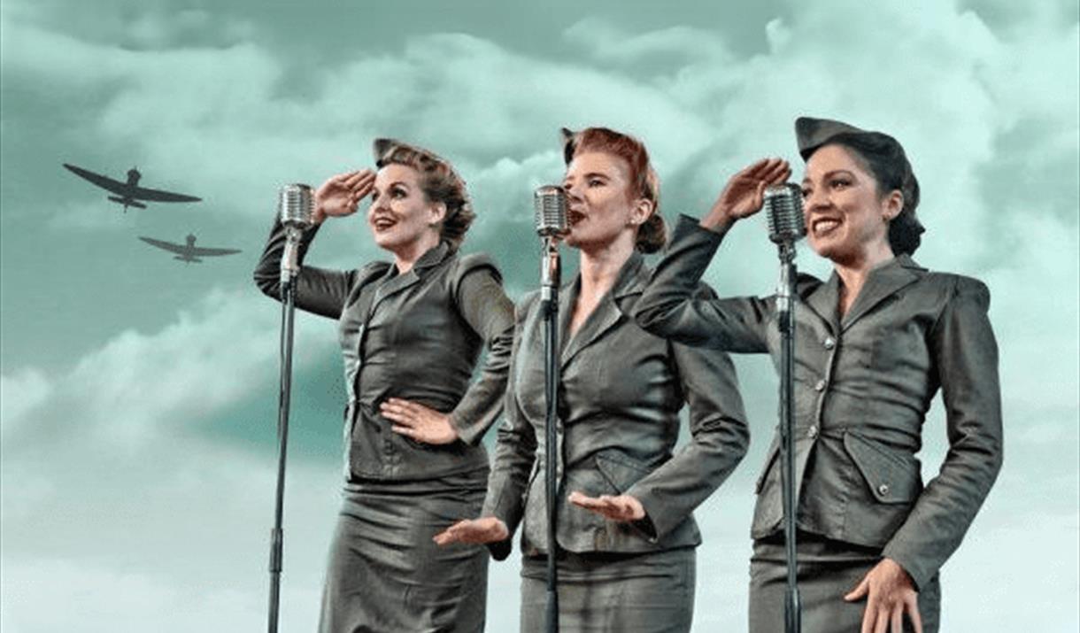 Three women in 1940's war time costume singing into microphones with 1940's war planes in the background