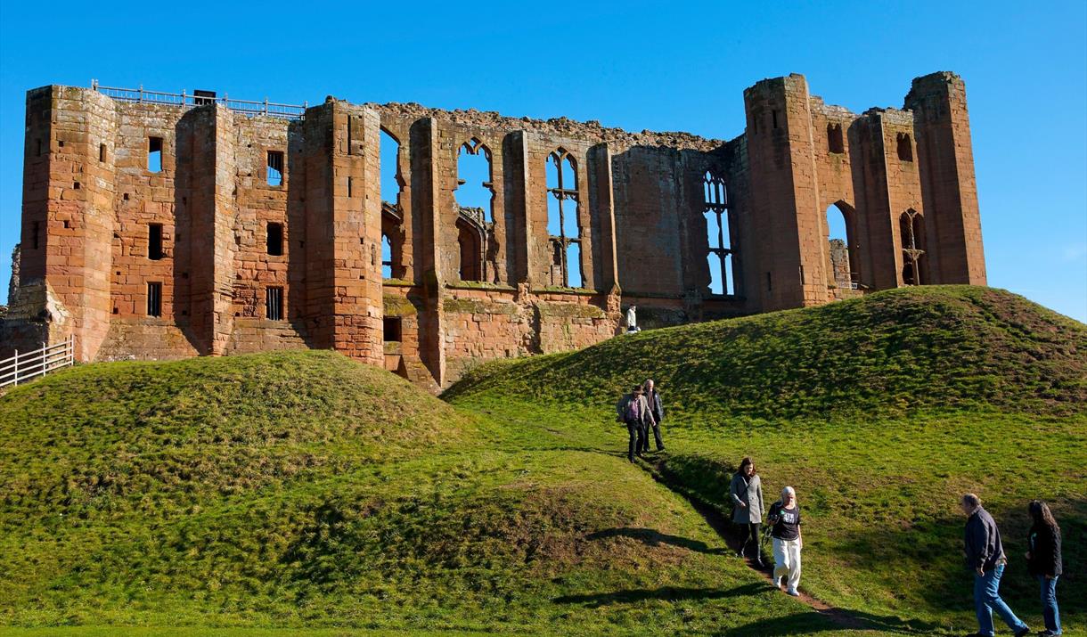 Looking up towards the ruins of Kenilworth Castle where people are climbing the slope towards it on a clear day with a bright blue sky in the backgrou
