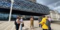 A group of five visitors stand in a circle listening to a tour guide who is wearing a bright yellow Roundhouse Birmingham coat. Birmingham Library rea