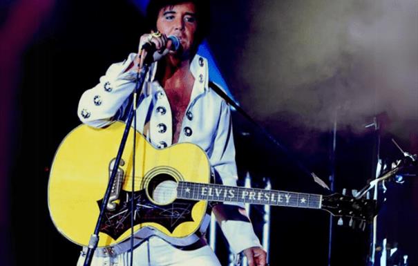 An Elvis tribute act in glittering white jumpsuit with a guitar and holding a microphone.
