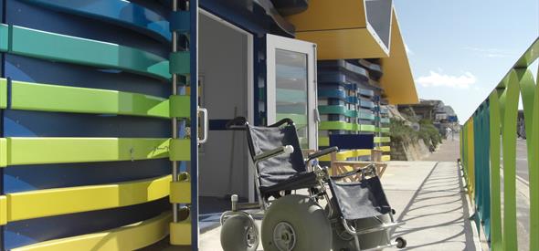 Beach wheelchait sat outside the green, blue and yellow accessible huts