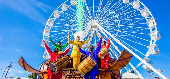 5 men playing instruments whilst dressed in colourful clothing in front of Bournemouths big wheel 