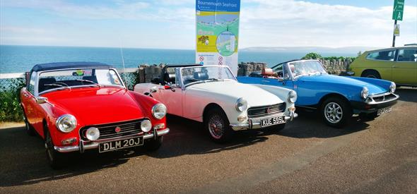 three classic cars in red, white and blue with the sea in the background