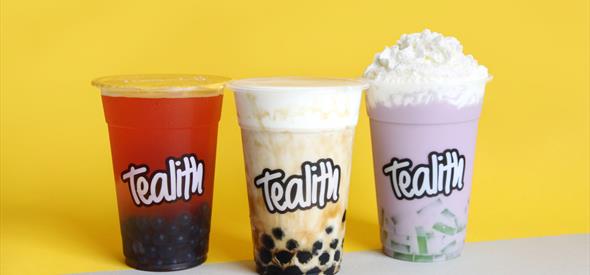 A shot of three bubble teas on a yellow background; one red, one creamy and one purple with foam.