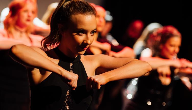 A close up of a young woman on a dark stage. She is lit with warm orange lighting and is in the middle of a dance. Her hair is brown and tied back in