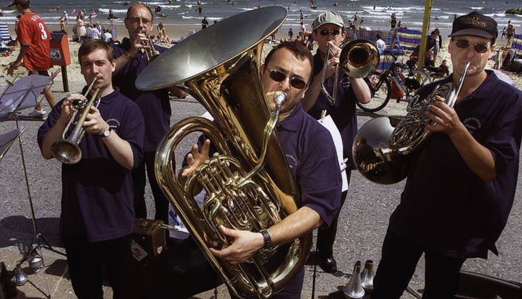 Musicians playing on the seafront prom with the sea and beach in the background and a tuba player centre foreground