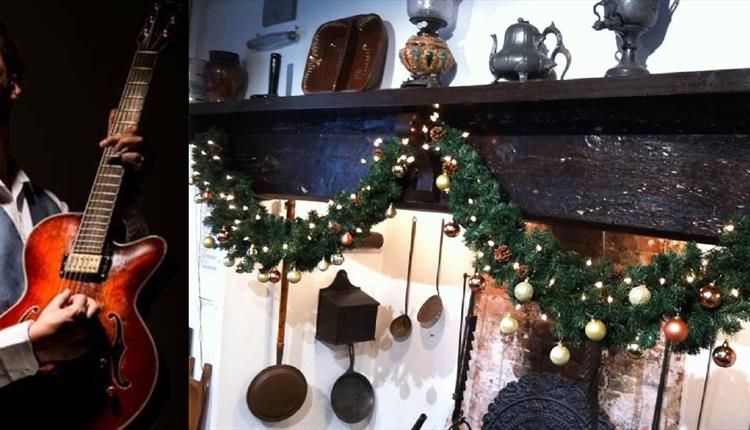 Ben standing in playing pose with guitar, wearing a waistcoat and black background, next to a Christmas decked fireplace