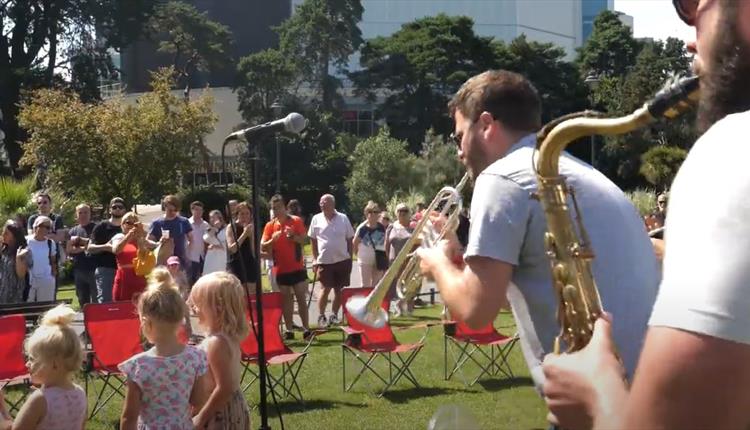 Outdoor concert with trumpet & saxophone players in foreground and children and parents in background, trees and grass