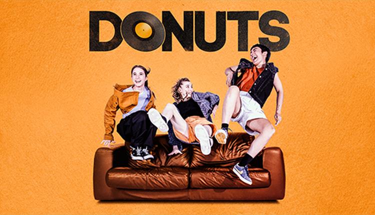 Three female dancers jump onto a brown sofa, laughing. The background is orange and text above them reads: DONUTS