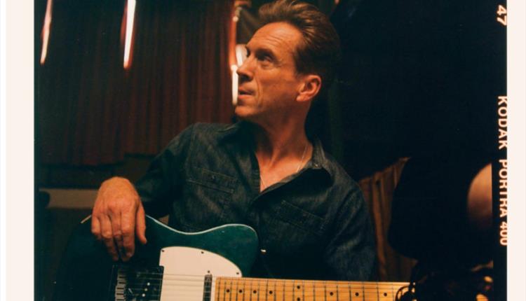 A side profile image of the artist Damian Lewis holding a guitar