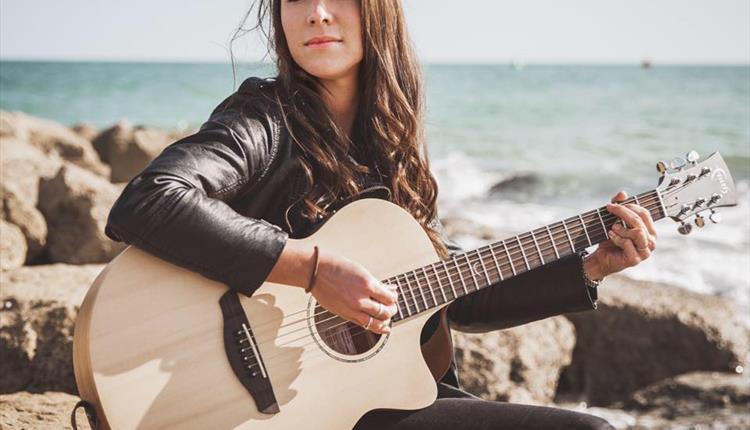 Gemma Leanne playing the guitar sitting on a rock by the sea