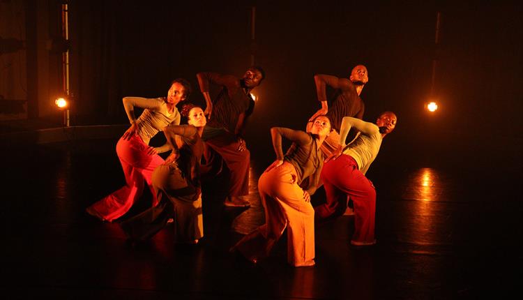 A photo from 'GROUNDATION - Dancing From the Archives' of a group of people on a dark stage, lit with warm orange light. They are all dressed in trous