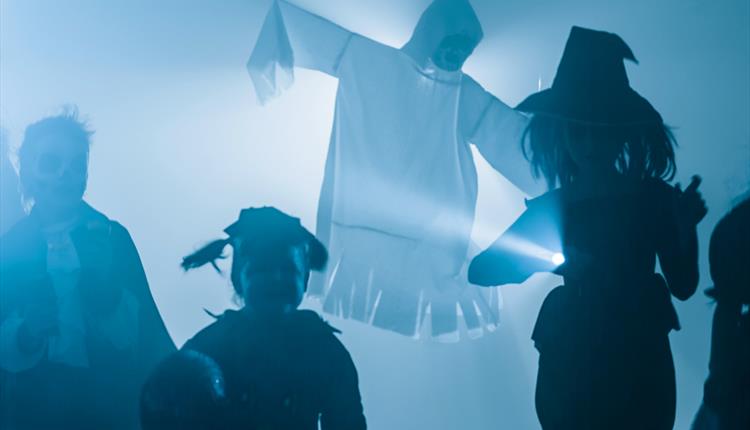 A spooky picture with a grey background and a pretend ghost decoration with the silhouettes of three children dressed in Halloween costume.