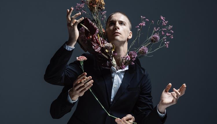 A promotional photo from the performance IMAGO. It shows a man stood in front of a dark blue/grey background. The man has a shaved head and is looking