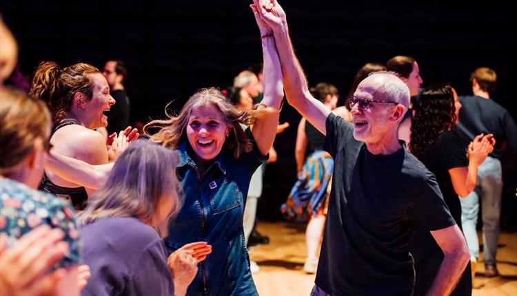 People joyfully dancing at a Ceilidh, with a man and woman smiling as they hold hands mid-dance, surrounded by a lively crowd in a warm, celebratory a