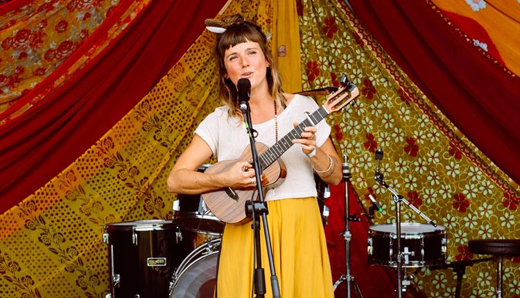 Female singer with ukulele standing in front of a red/orange backdrop with a drum kit behind her.