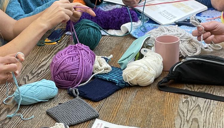 A table cluttered with yarn and instructions, several pairs of hands each work on a different coloured knit or crochet.