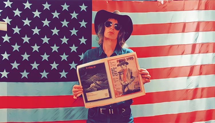 Mike scott posing in cowboy hat and sunglasses, looking towards camera holding an open newspaper with American flag as backdrop