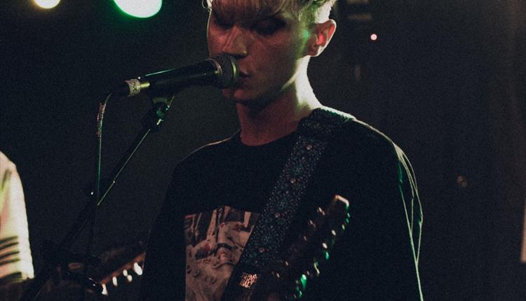 Young blond guitarist standing in front of a microphone singing. Very dark background and low lighting