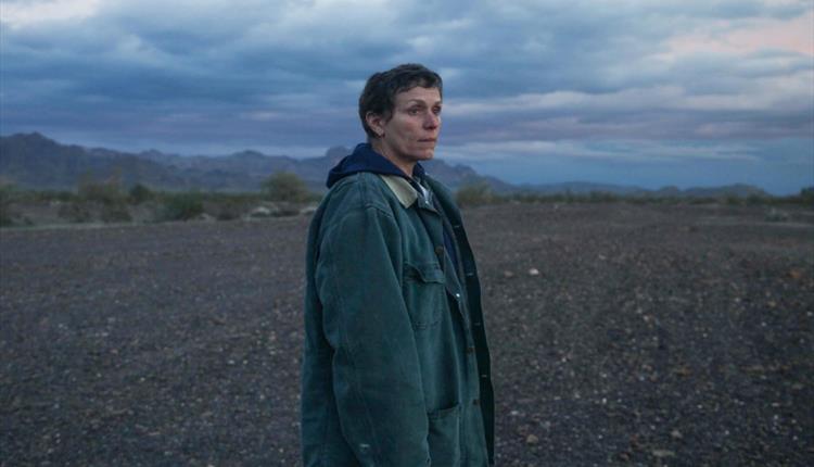 A woman wearing short hair and a baggy coat stands on an empty patch of land, staring into the distance.