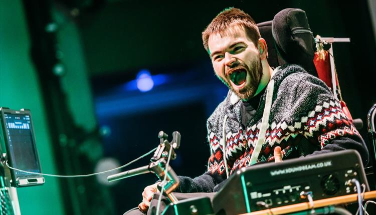 A photo of a young man on a stage lit with blue and green lights. He is in a wheelchair with a microphone set up in front of him. He has short brown h