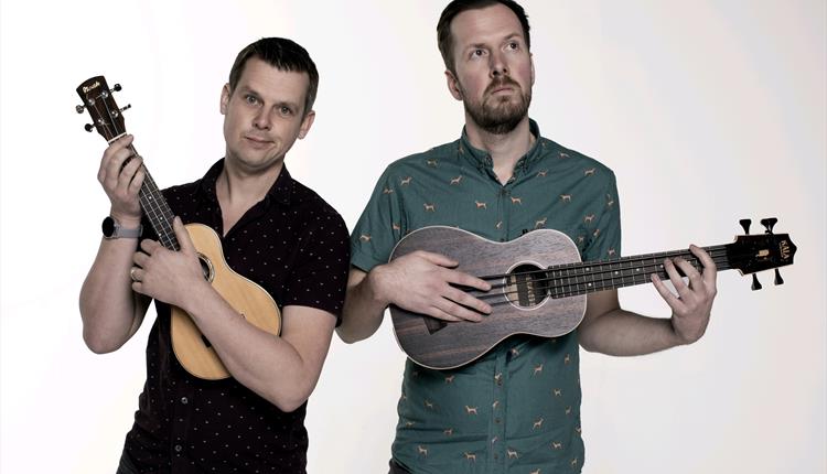 Tow men playing ukuleles facing front one smaller than the other both with quizical expressions on their faces