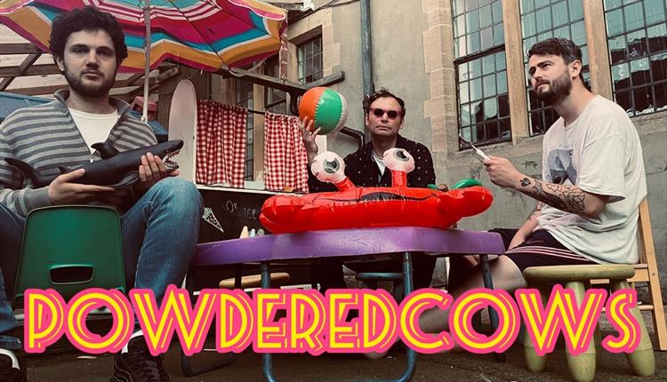 3 men sitting around apurpl table with a red inflatable on it holding instruments and the words powderedcow in front of them.