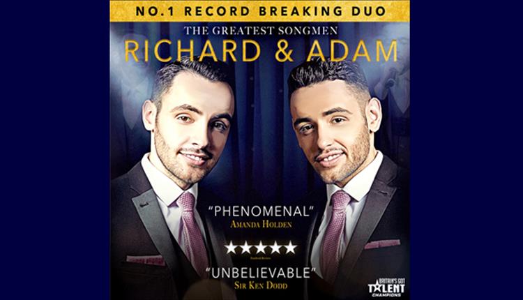 Richard and Adam in black suits and lilac ties, smiling at camera with dark curtain background