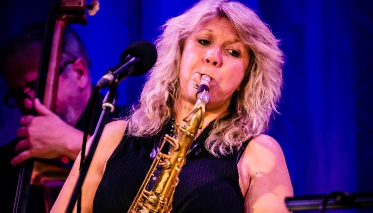 Blond female saxophonist in front of a microphone with a male double bass player in the background