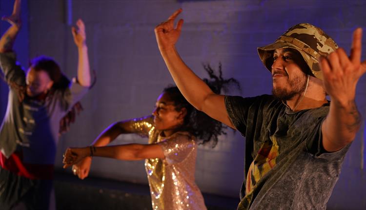 The top halves of three dancers in a night club setting. Two males are either side of a female, each in their mid-20s and of dual heritage.