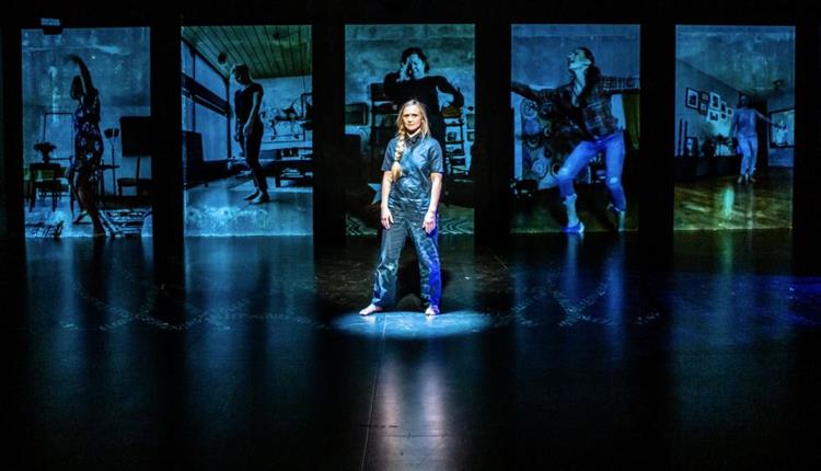 A photo from 'When The Bleeding Stops' of a woman on a dark stage. She is lit in a blue light and behind her there is blue projections of many differe