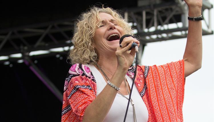 Female singer with blond curly hair singing into a microphone, wearing a red jacket and with her left hand up in the air