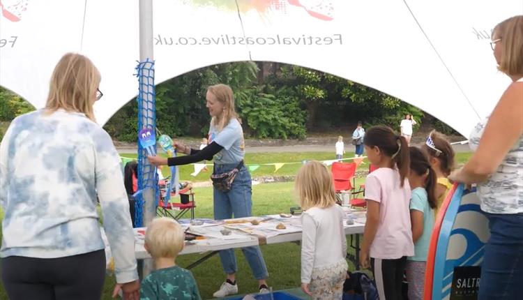 Children and adults in front of trestle table with arts and crafts materials under a stretch tent  - background of trees and shrubs