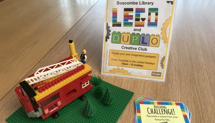 A red fire engine made from lego bricks on a green base. To the right are some brick building challenge cards and behind is a poster saying "Lego and