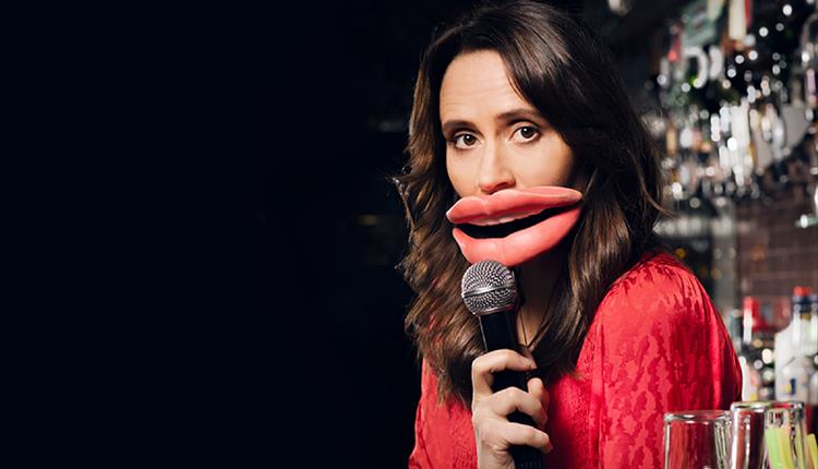 Nina Conti looking at camera speaking into microphone with huge red lip props wearing a red blouse. bar background.