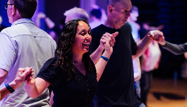 A photo from a Ceilidh dance. It features a man and a woman stood among many other people dancing in a big, dark hall. The hall is lit with purple lig