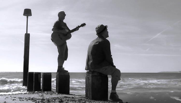 Black & white image of two men, one sitting on a breakwater and the other standing on the breakwater playing a guitar, looking out to sea