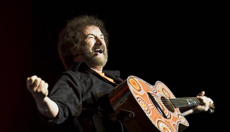 Kevin Bloody Wilson, Kevin Wilson on stage holding a guitar with strained expression on his face with dark background