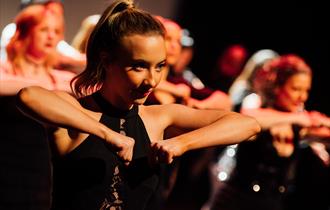 A close up of a young woman on a dark stage. She is lit with warm orange lighting and is in the middle of a dance. Her hair is brown and tied back in