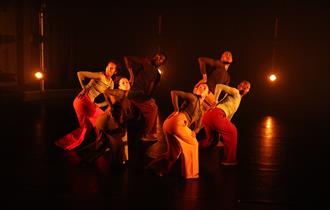 A photo from 'GROUNDATION - Dancing From the Archives' of a group of people on a dark stage, lit with warm orange light. They are all dressed in trous