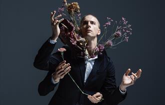 A promotional photo from the performance IMAGO. It shows a man stood in front of a dark blue/grey background. The man has a shaved head and is looking