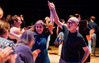 People joyfully dancing at a Ceilidh, with a man and woman smiling as they hold hands mid-dance, surrounded by a lively crowd in a warm, celebratory a