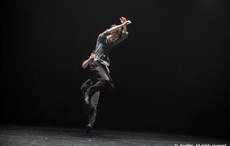 A photo of Kontemporary Korea. Two men on a black stage. Both wearing black trousers and socks and a charcoal grey, loose, long sleeved top. One man i