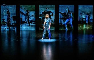 A photo from 'When The Bleeding Stops' of a woman on a dark stage. She is lit in a blue light and behind her there is blue projections of many differe