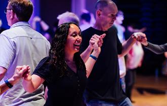 A photo from a Ceilidh dance. It features a man and a woman stood among many other people dancing in a big, dark hall. The hall is lit with purple lig